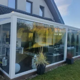 Szklana zabudowa tarasu przylegająca do domu, widoczne meble ogrodowe wewnątrz, dekoracyjne donice z trawami na zewnątrz.