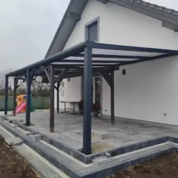 Zadaszona konstrukcja z elementami drewnianymi i metalowymi, przylegająca do białego domu z szarą dachówką, z widocznym fragmentem tarasu z szarej kostki brukowej i placem zabaw w tle.