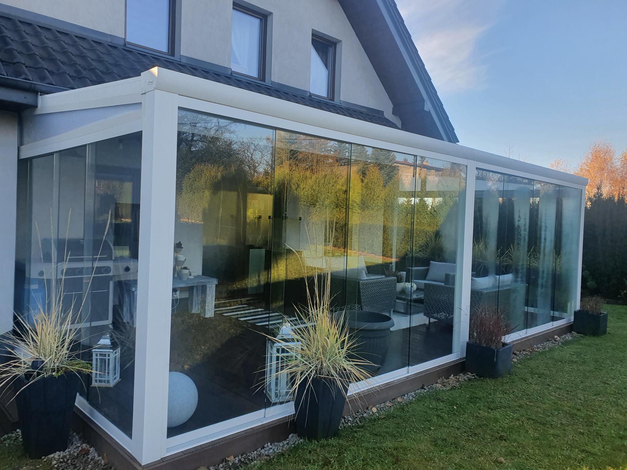 Szklana zabudowa tarasu przylegająca do domu, widoczne meble ogrodowe wewnątrz, dekoracyjne donice z trawami na zewnątrz.