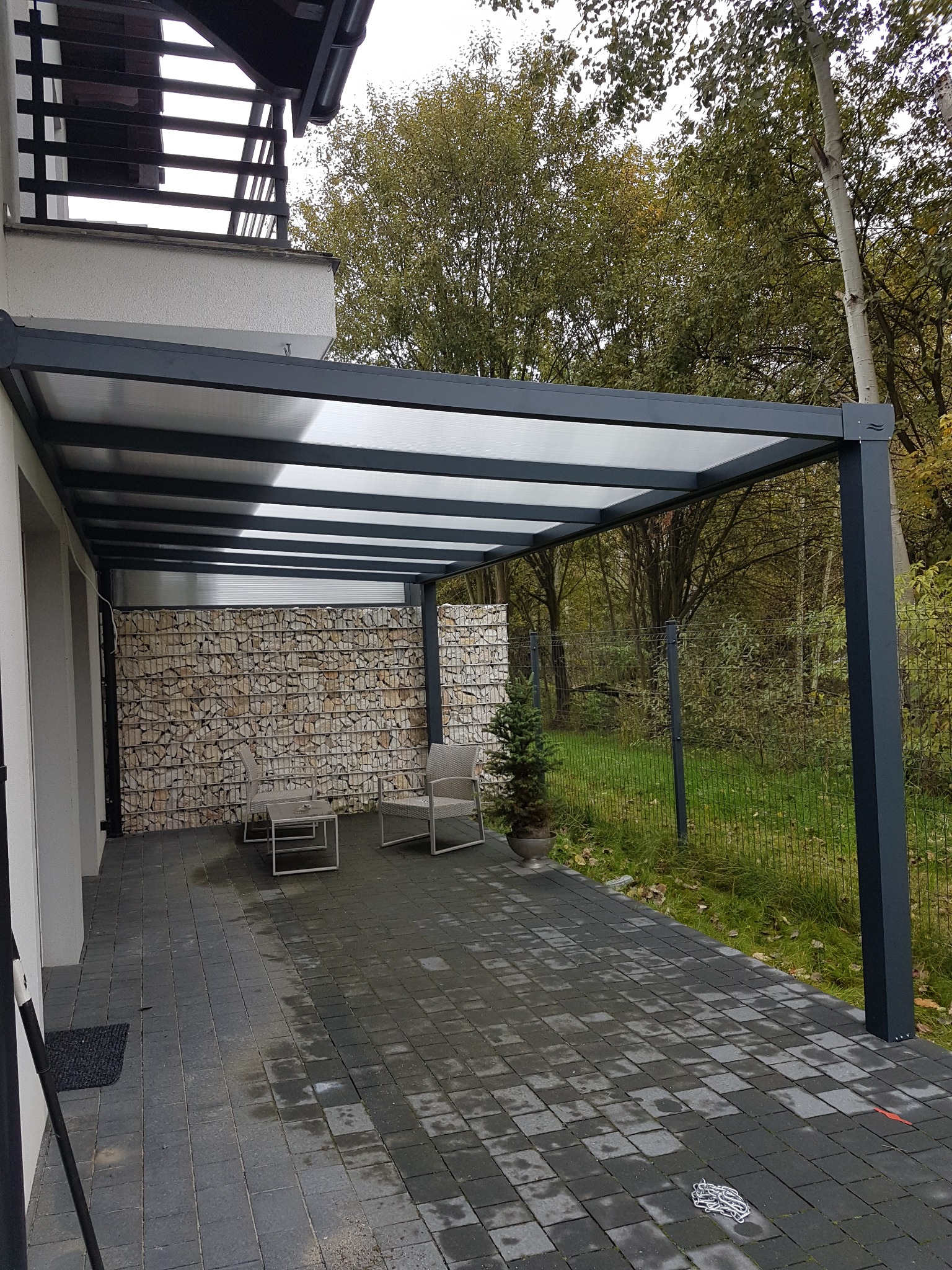 Nowoczesna, grafitowa pergola z przezroczystym zadaszeniem, przylegająca do budynku, z gabionową ścianą w tle i dwoma krzesłami ogrodowymi.