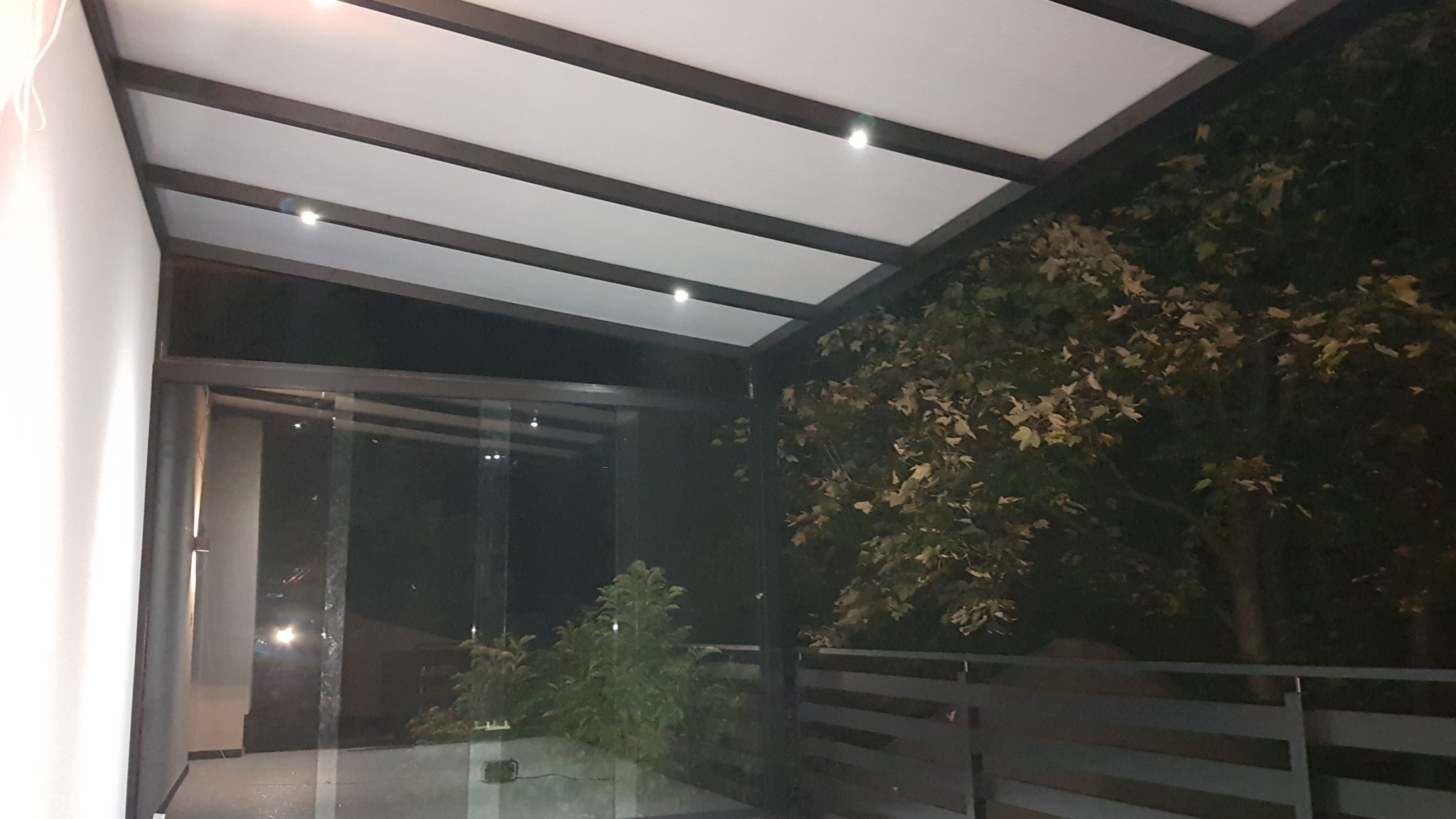 Nowoczesna pergola z białym zadaszeniem i oświetleniem punktowym, widok z zewnątrz na drzewo z opadającymi liśćmi i szarą balustradę.