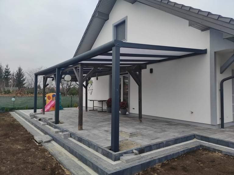 Zadaszona konstrukcja z elementami drewnianymi i metalowymi, przylegająca do białego domu z szarą dachówką, z widocznym fragmentem tarasu z szarej kostki brukowej i placem zabaw w tle.