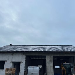 PROSTE DACHY USŁUGI CIESIELSKO-DEKARSKIE MARCIN KWIETNIEWSKI - Surowy budynek z ciemną dachówką w trakcie budowy, widoczny mini-koparka i palety. Dach pokryty delikatnym śniegiem, pochmurne niebo w tle.