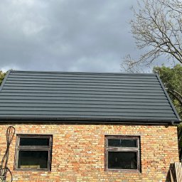 PROSTE DACHY USŁUGI CIESIELSKO-DEKARSKIE MARCIN KWIETNIEWSKI - Szary dach z blachy na ceglanym budynku z dwoma oknami, widok z dołu, pochmurne niebo w tle. Widoczne drzewa i fragment samochodu.