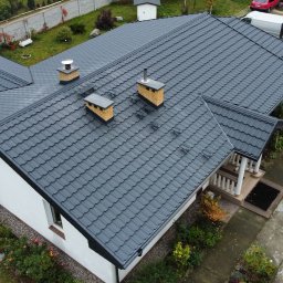 PROSTE DACHY USŁUGI CIESIELSKO-DEKARSKIE MARCIN KWIETNIEWSKI - Antracytowy dach z blachodachówki modułowej na białym domu jednorodzinnym, widok z góry. Na dachu trzy kominy z ceglanymi elementami. Zadbane otoczenie domu.