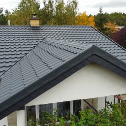 PROSTE DACHY USŁUGI CIESIELSKO-DEKARSKIE MARCIN KWIETNIEWSKI - Grafitowy dach z blachodachówki modułowej na domu jednorodzinnym, widok z góry, komin z cegły klinkierowej, jesienne drzewa w tle, widoczne rusztowanie.