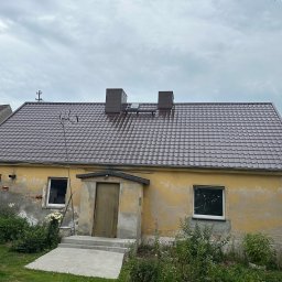 PROSTE DACHY USŁUGI CIESIELSKO-DEKARSKIE MARCIN KWIETNIEWSKI - Dom z nowym, brązowym dachem z blachy trapezowej i dwoma kominami. Widoczne ślady napraw na elewacji. Pochmurne niebo w tle.