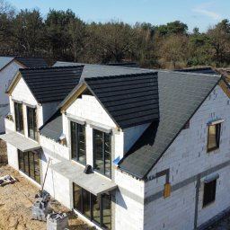 PROSTE DACHY USŁUGI CIESIELSKO-DEKARSKIE MARCIN KWIETNIEWSKI - Nowoczesne domy w trakcie budowy z ciemnymi dachami, widoczne fundamenty kolejnych budynków i ekipa budowlana. Ujęcie z góry, słoneczny dzień.