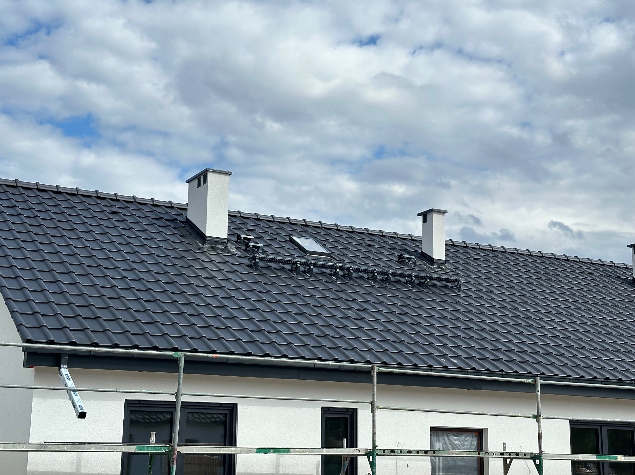Nowy, grafitowy dach z dachówki ceramicznej z oknem dachowym i systemem odśnieżania, białe kominy, elewacja w trakcie prac wykończeniowych, widoczne rusztowanie.