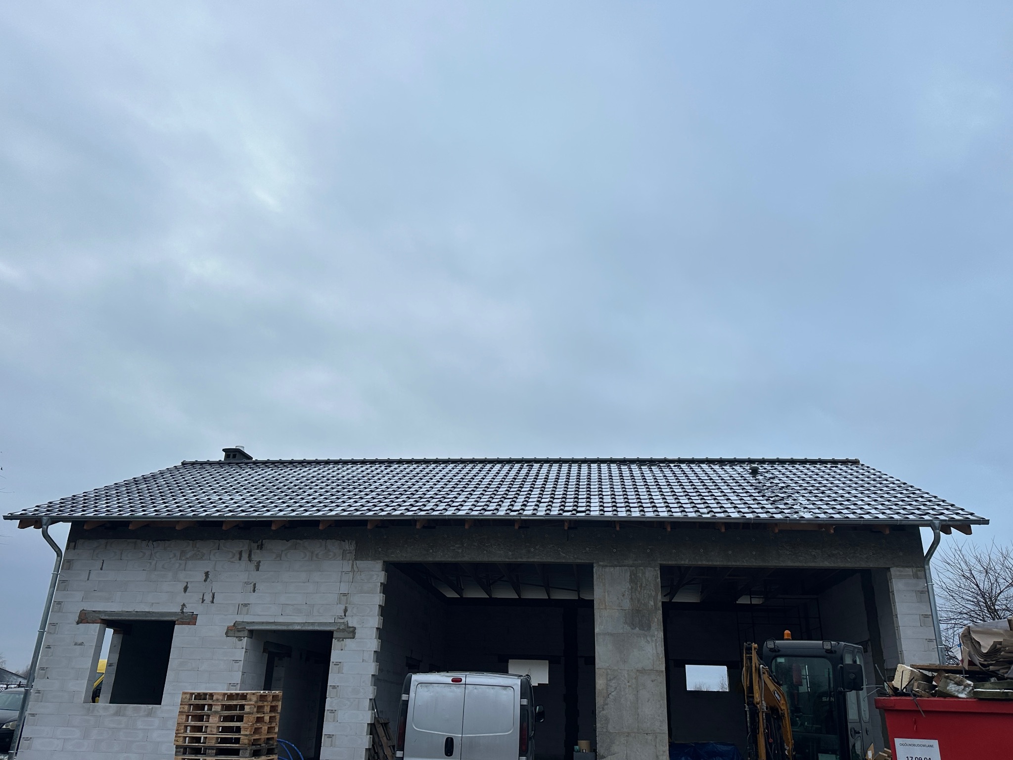 Surowy budynek z ciemną dachówką w trakcie budowy, widoczny mini-koparka i palety. Dach pokryty delikatnym śniegiem, pochmurne niebo w tle.