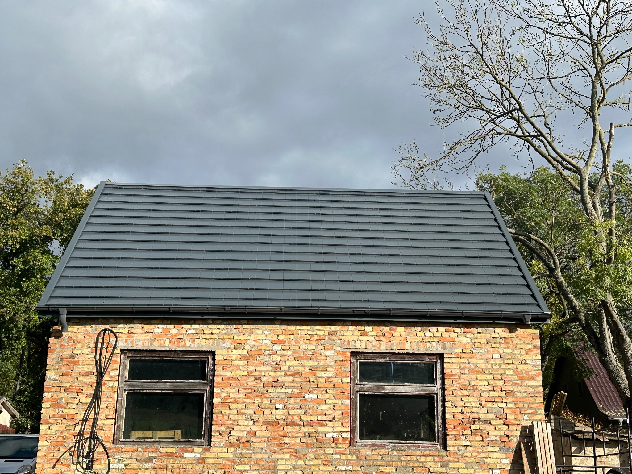 Szary dach z blachy na ceglanym budynku z dwoma oknami, widok z dołu, pochmurne niebo w tle. Widoczne drzewa i fragment samochodu.