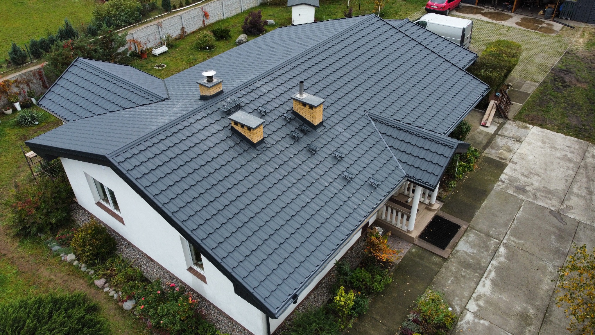 Antracytowy dach z blachodachówki modułowej na białym domu jednorodzinnym, widok z góry. Na dachu trzy kominy z ceglanymi elementami. Zadbane otoczenie domu.
