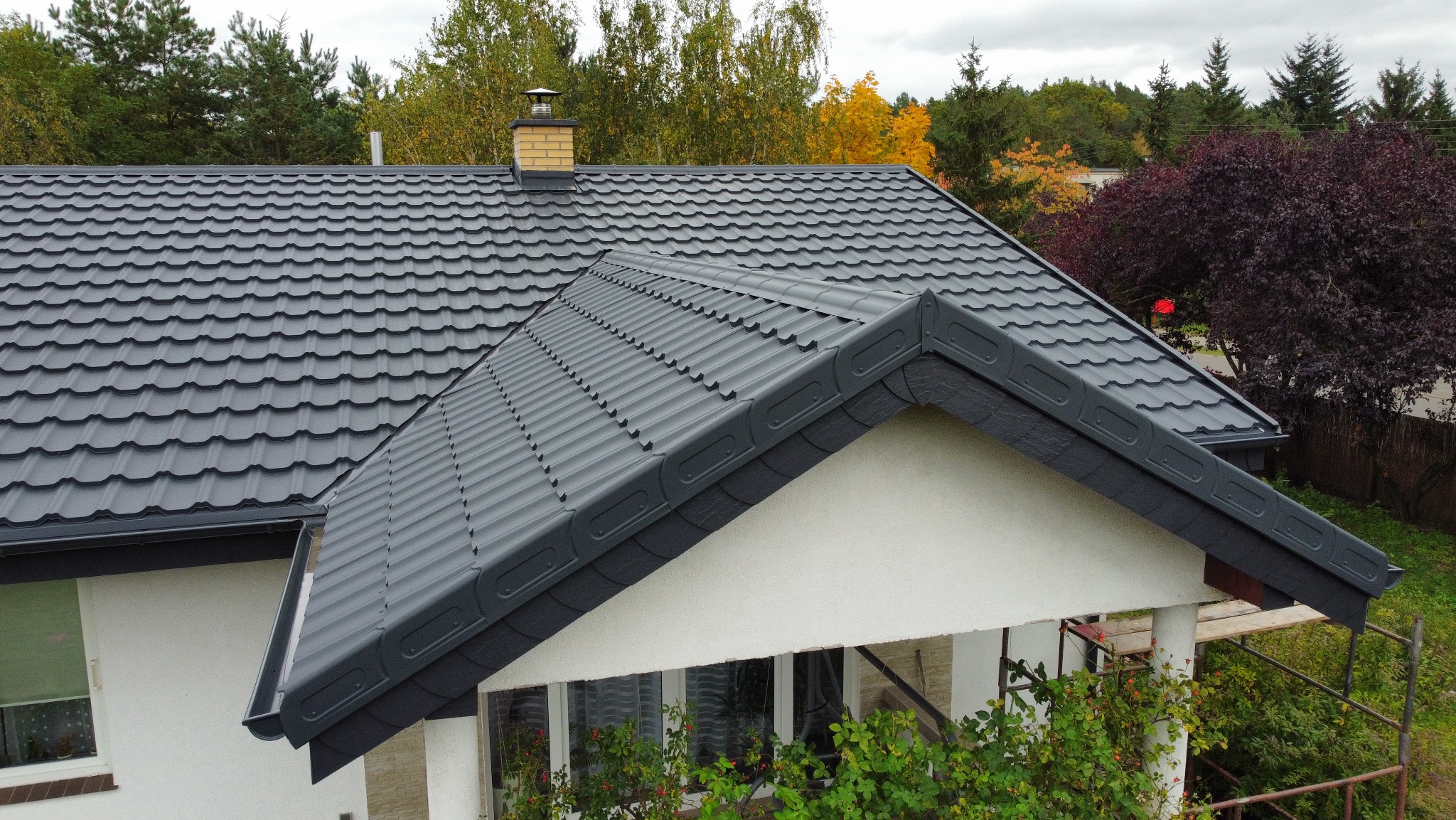 Grafitowy dach z blachodachówki modułowej na domu jednorodzinnym, widok z góry, komin z cegły klinkierowej, jesienne drzewa w tle, widoczne rusztowanie.