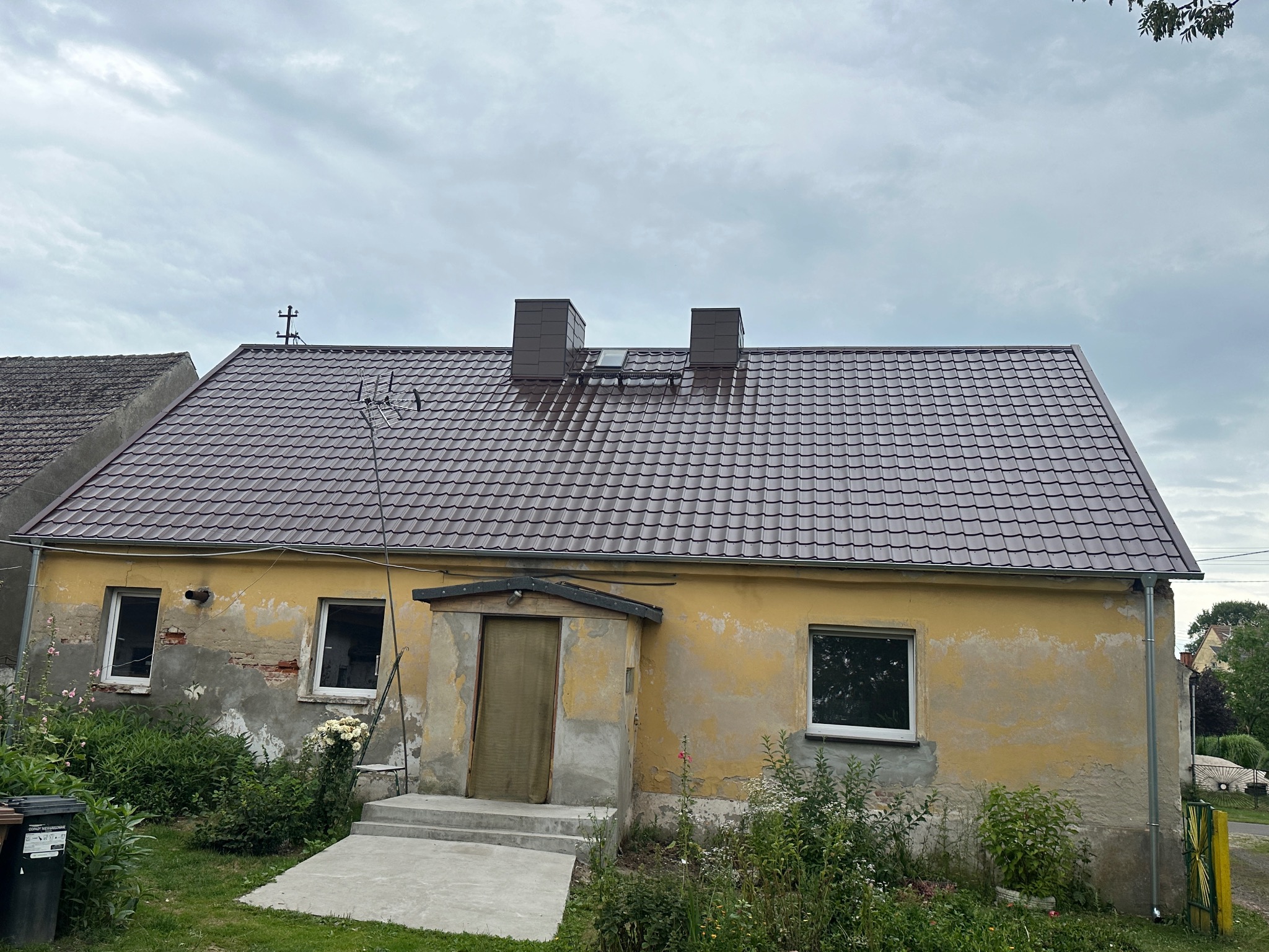 Dom z nowym, brązowym dachem z blachy trapezowej i dwoma kominami. Widoczne ślady napraw na elewacji. Pochmurne niebo w tle.