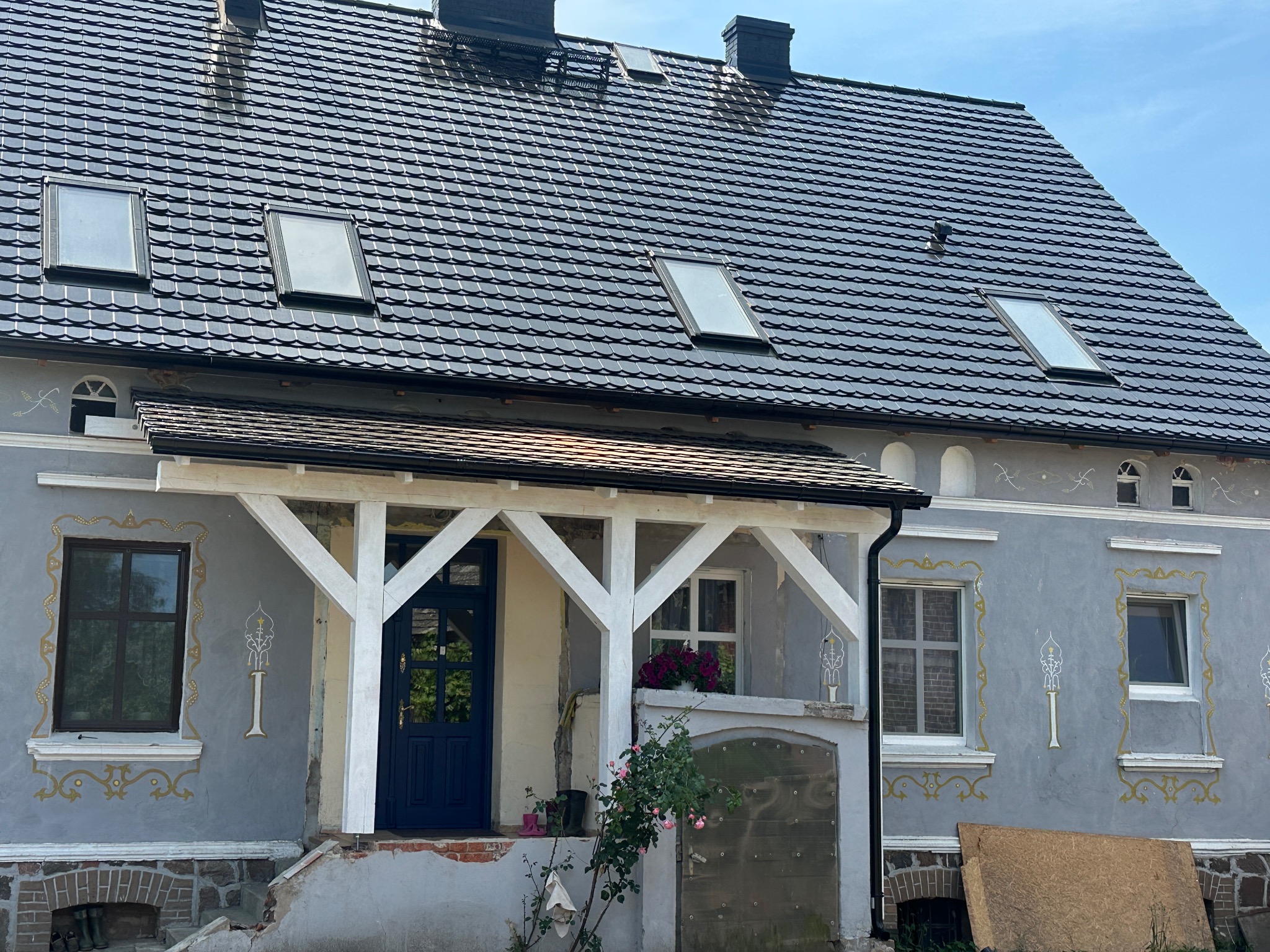 Dom z ciemnoszarym dachem z dachówki ceramicznej i oknami dachowymi. Widoczny fragment elewacji z dekoracjami i weranda z białymi elementami konstrukcyjnymi.