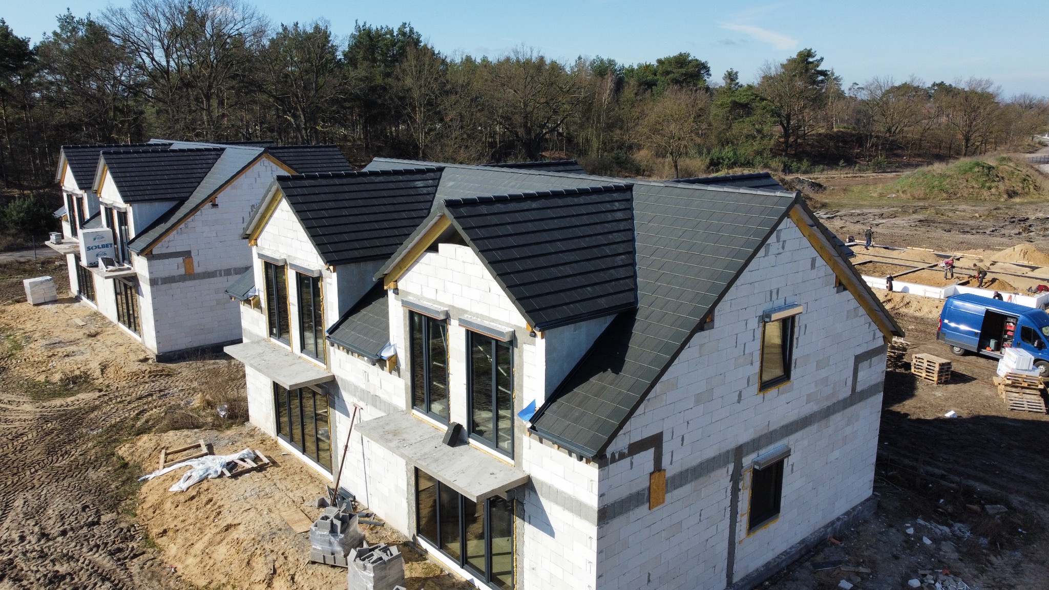 Nowoczesne domy w trakcie budowy z ciemnymi dachami, widoczne fundamenty kolejnych budynków i ekipa budowlana. Ujęcie z góry, słoneczny dzień.