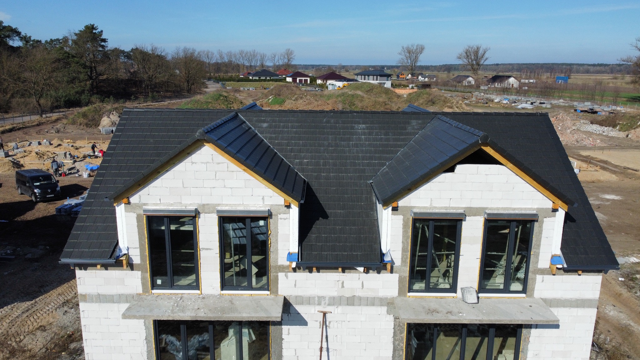 Dom w trakcie budowy z ciemnoszarą dachówką, widok z góry. Na pierwszym planie okna z czarnymi ramami. W tle zabudowa jednorodzinna i teren budowy.