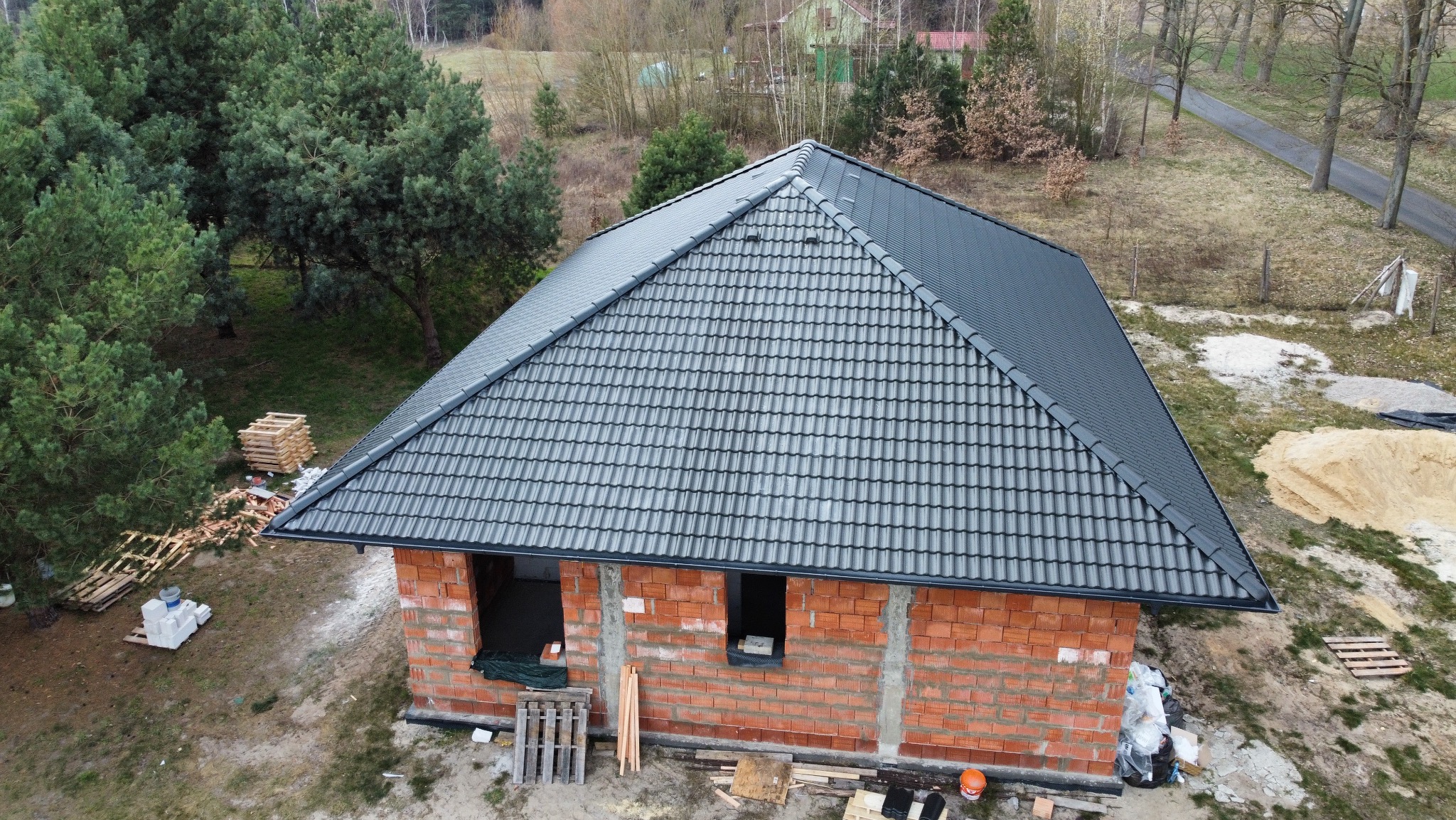 Nowoczesny, grafitowy dach z dachówki ceramicznej na surowym budynku z cegły. Widoczne rynny i obróbki blacharskie. Ujęcie z drona, zieleń w tle.