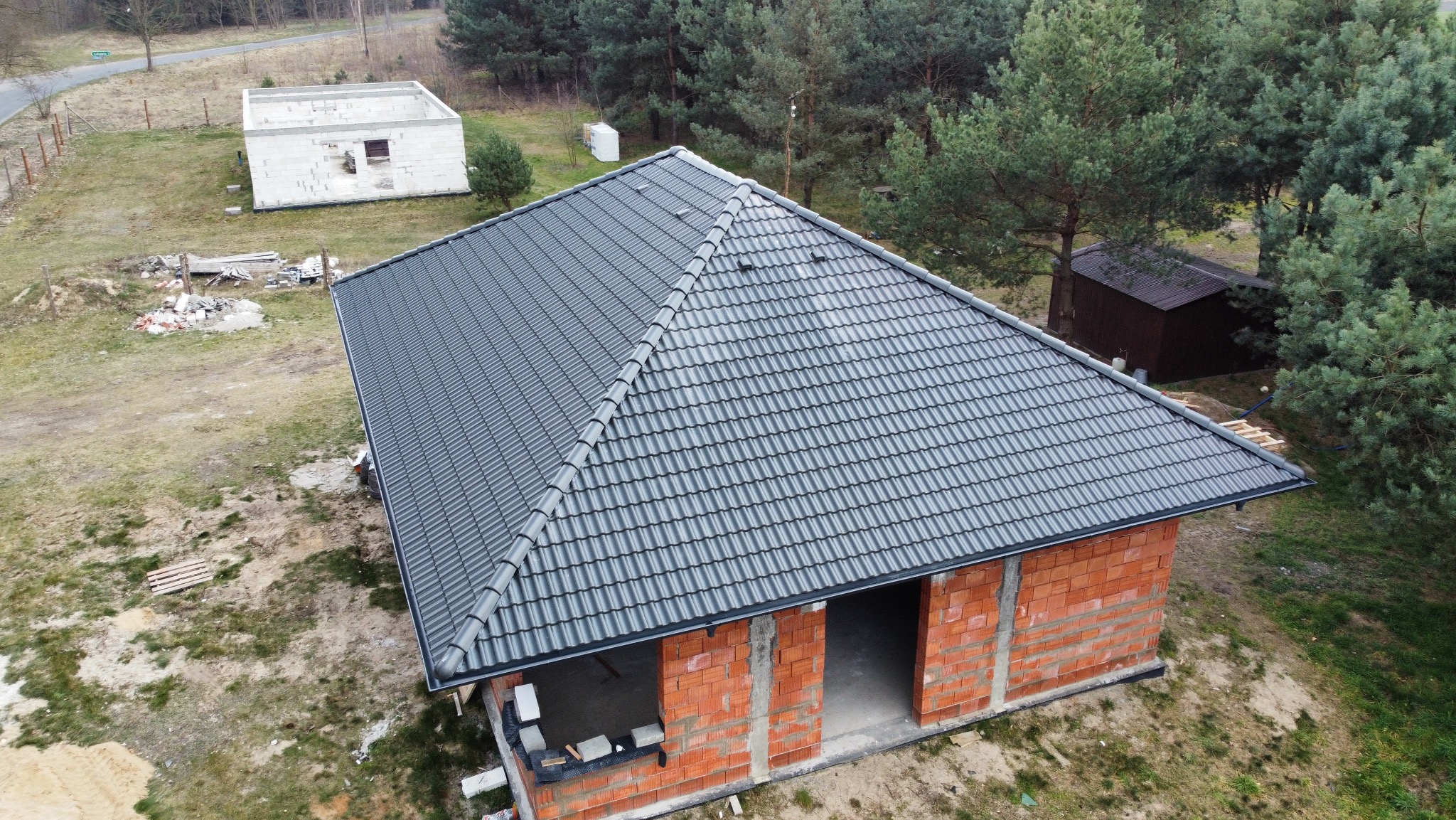 Nowy, grafitowy dach z dachówki ceramicznej na surowym budynku z czerwonej cegły. Widok z góry ukazuje precyzyjne ułożenie dachówek i otoczenie z drzewami.