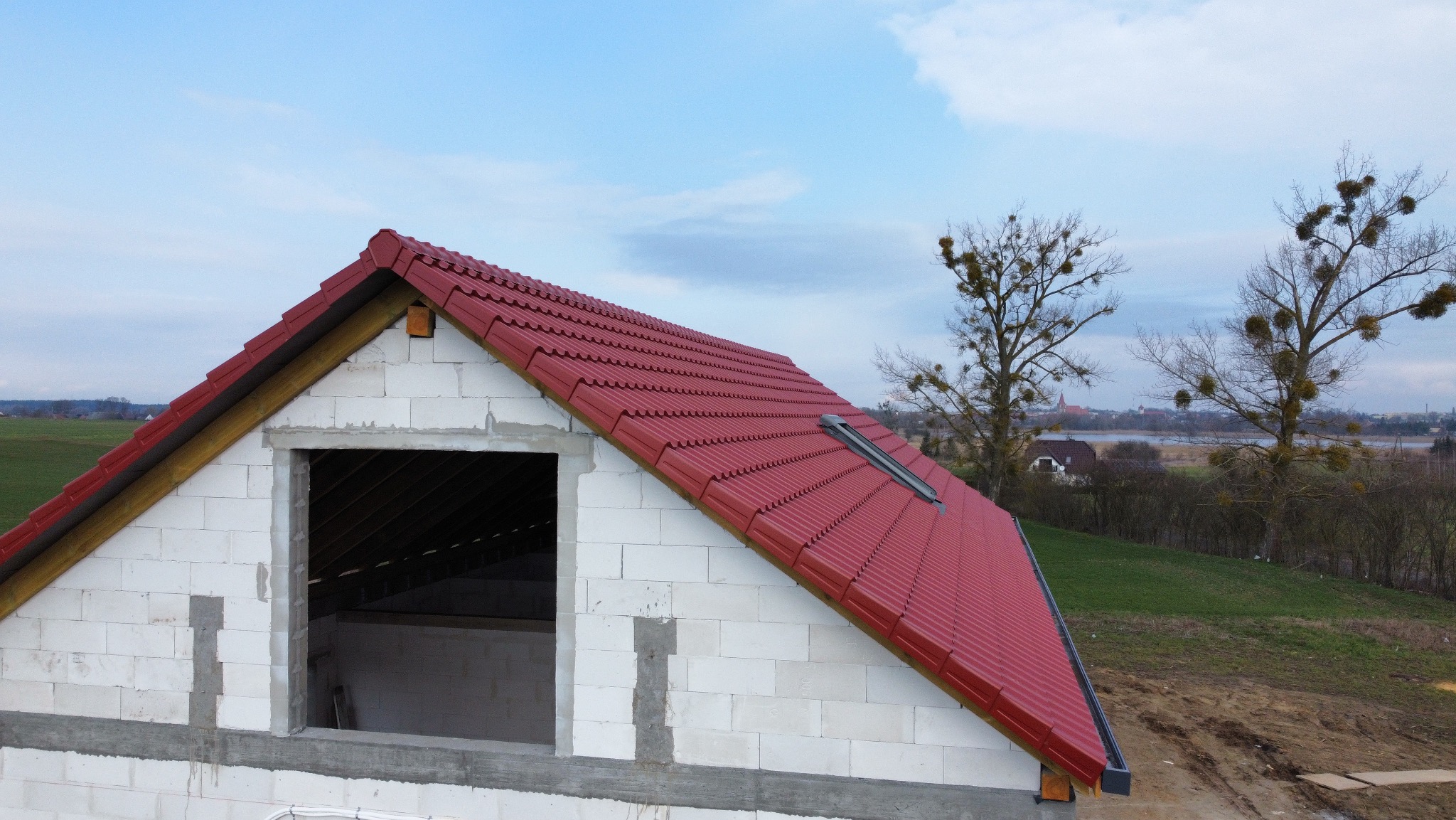 Czerwona dachówka na surowym budynku z pustaków, widok z góry. Okno dachowe i otwarty otwór okienny. W tle drzewa i krajobraz.