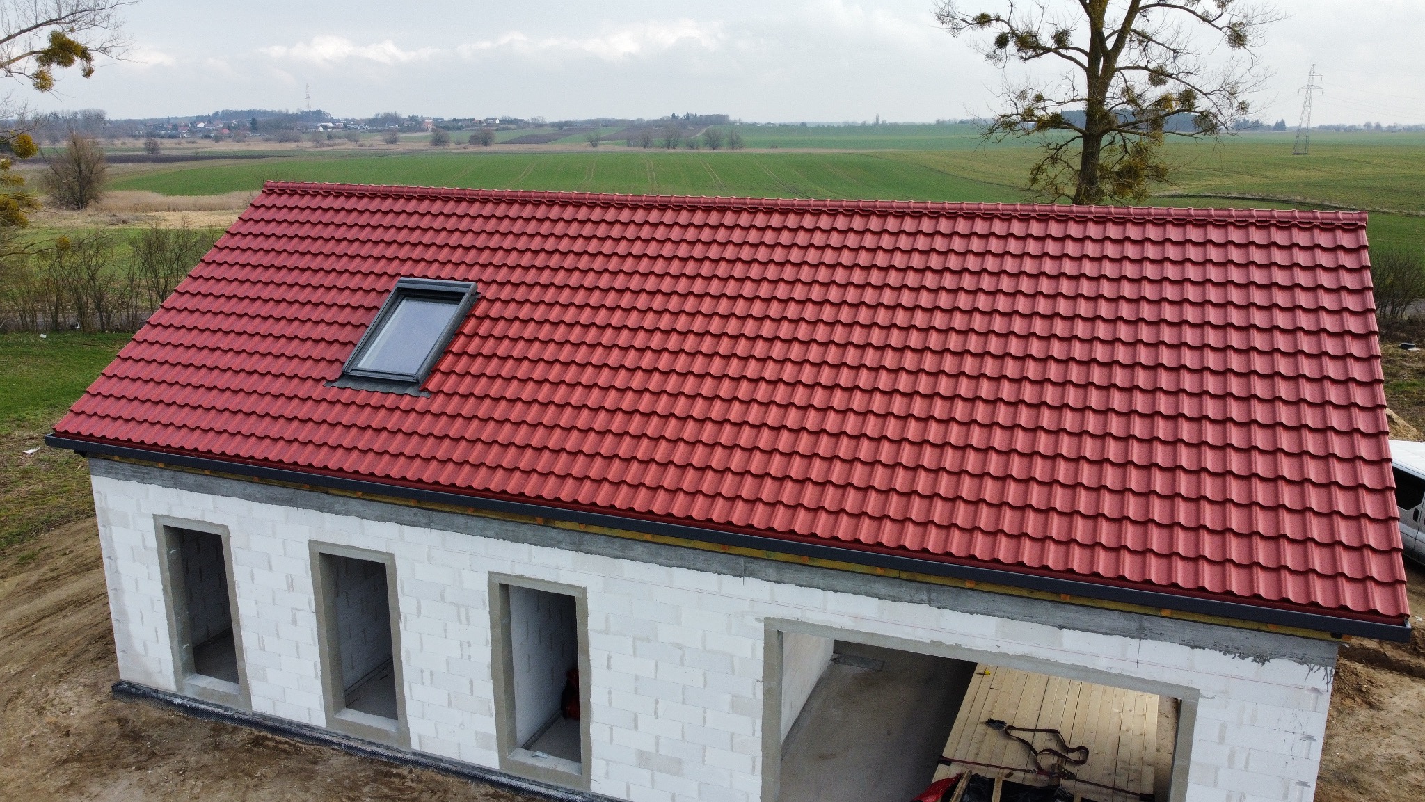 Czerwony dach z dachówką ceramiczną i oknem połaciowym na surowym budynku z betonu komórkowego, na tle zielonych pól i drzew.