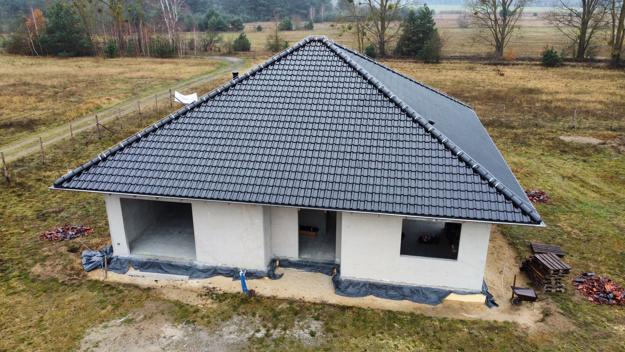 Nowoczesny dom w trakcie budowy z czarną dachówką karpiówką, widok z góry. Surowy stan, brak okien i drzwi. W tle łąka i las. Dachówka ułożona równo.