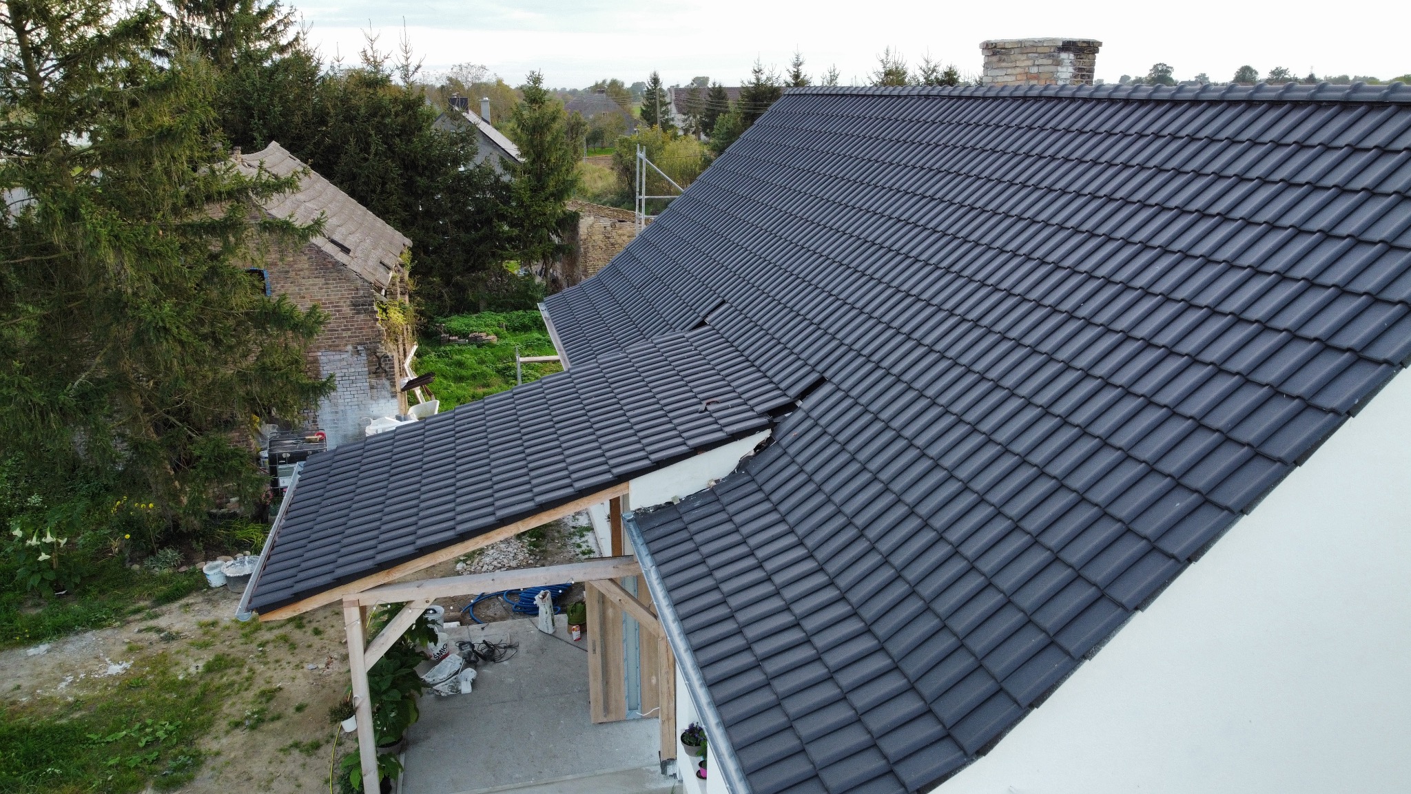 Ciemnoszary dach z dachówki ceramicznej, widok z góry, z fragmentem drewnianej konstrukcji werandy i otoczeniem zieleni. Nowoczesne pokrycie dachowe na tle wiejskiego krajobrazu.