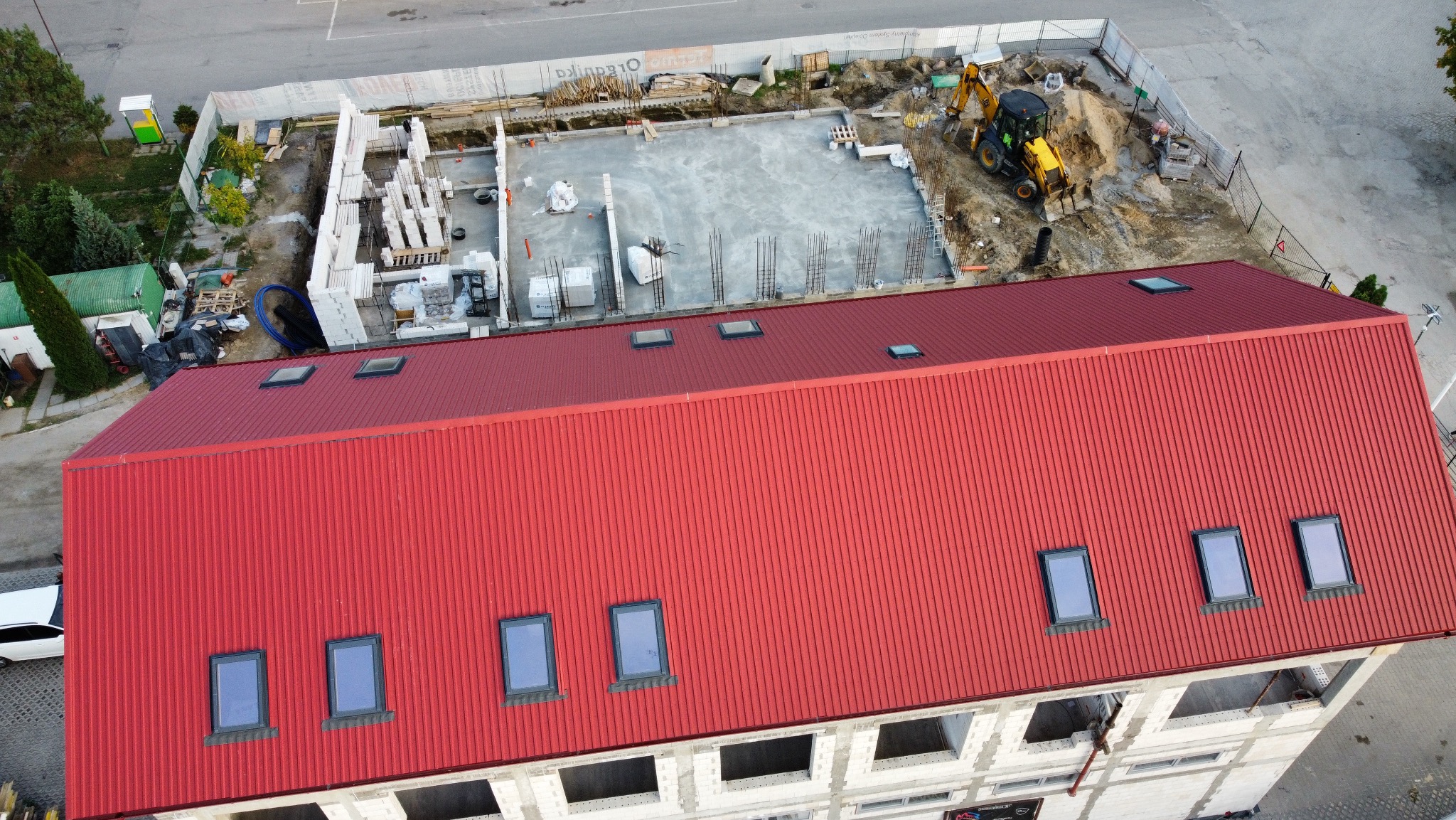 Czerwony dach z oknami połaciowymi na budynku w trakcie budowy, widok z góry. W tle plac budowy z fundamentami i sprzętem budowlanym.