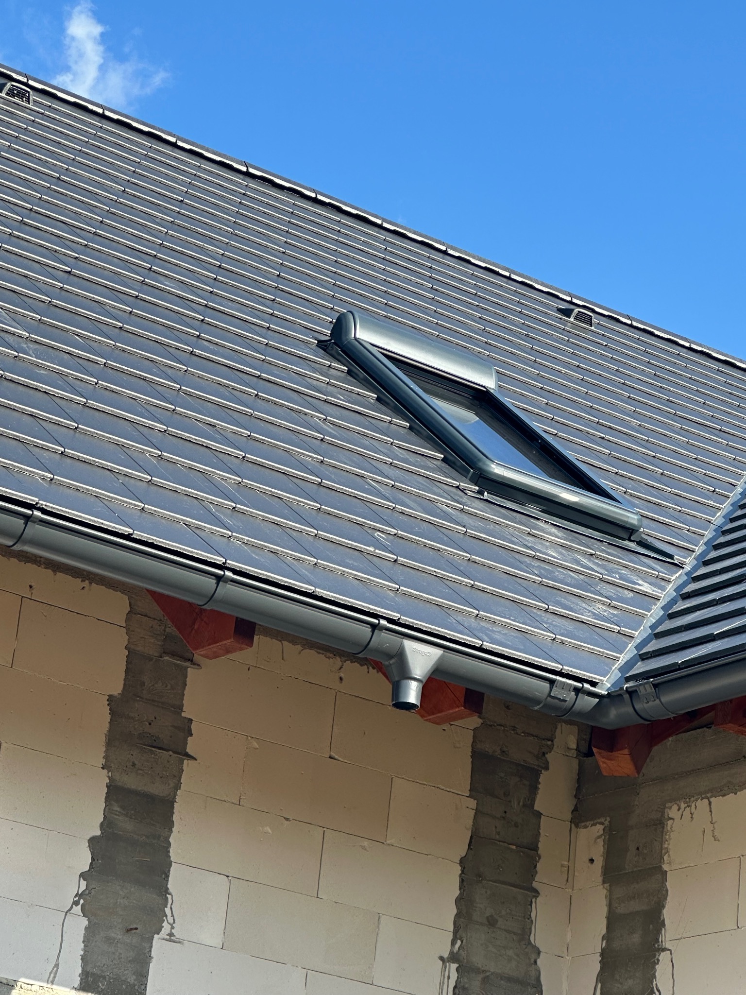 Grafitowy dach z dachówki ceramicznej, okno dachowe i rynna w kolorze antracytu na tle błękitnego nieba. Widoczne fragmenty ściany budynku w trakcie budowy.