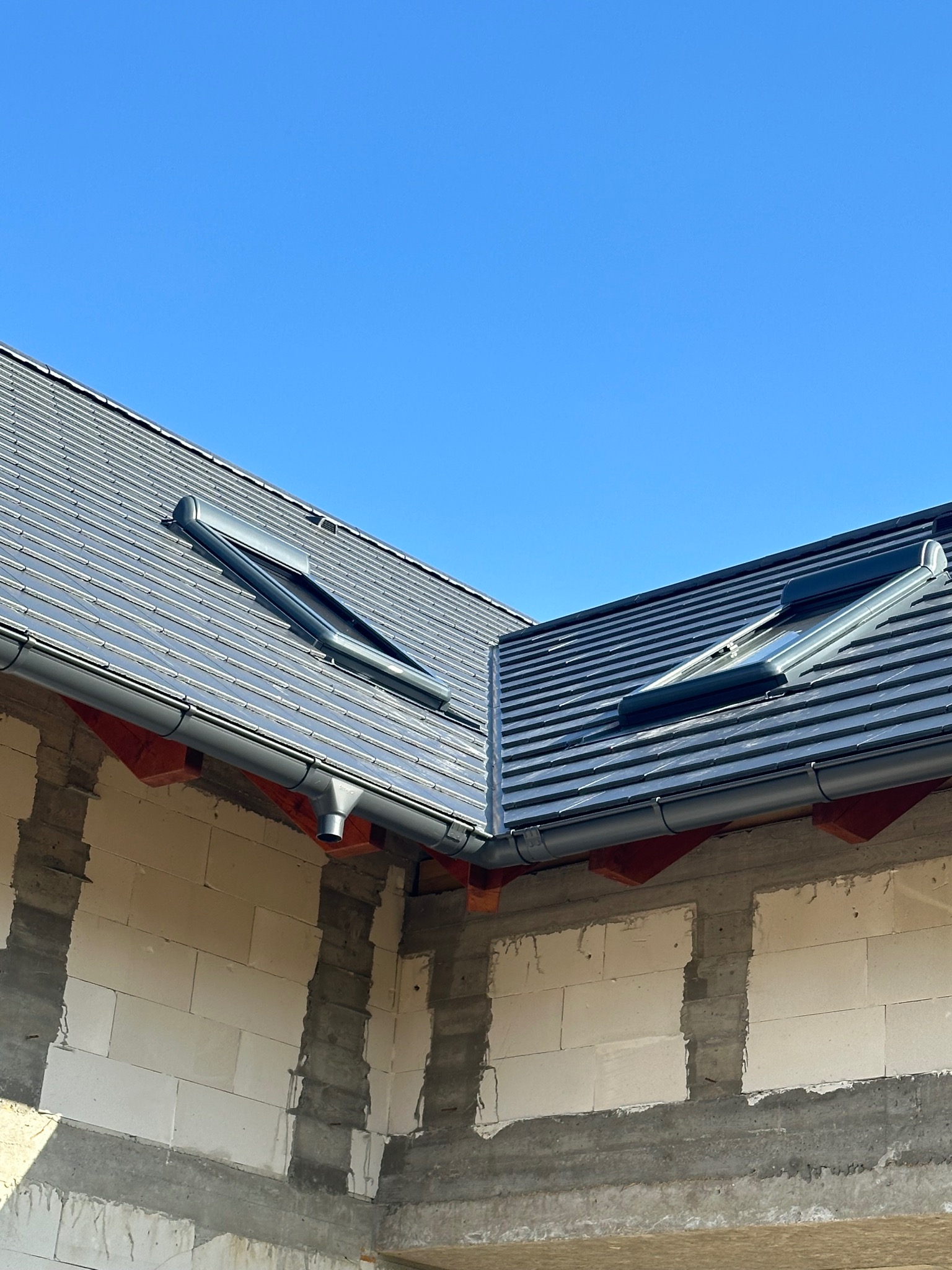 Szary dach z dachówką ceramiczną i oknami dachowymi, widoczny fragment ściany budynku z białego betonu komórkowego i szara rynna. Błękitne niebo w tle.