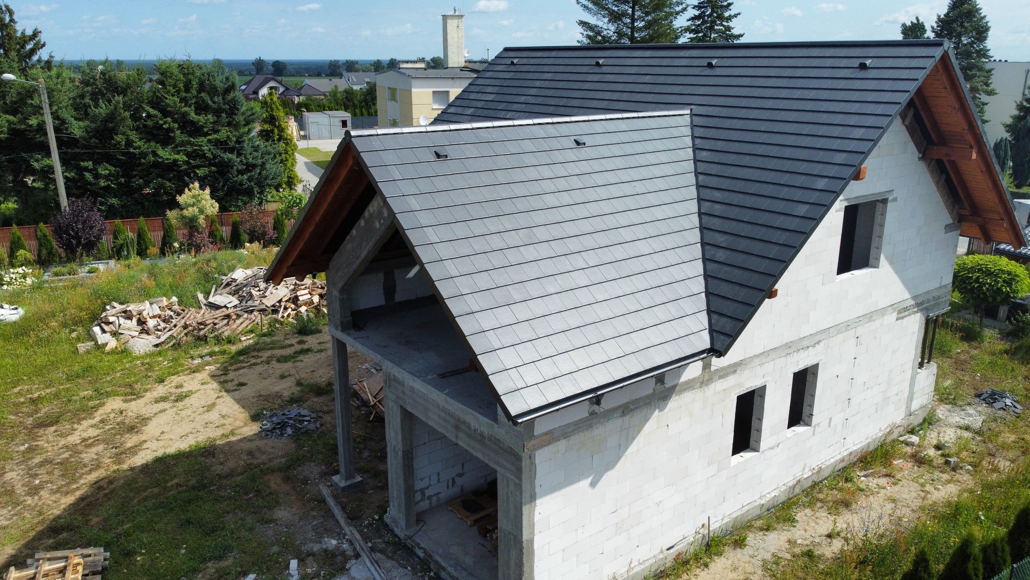 Dom w trakcie budowy z częściowo ukończonym dachem pokrytym szarą dachówką. Widoczne niezabudowane okna i elementy konstrukcyjne. W tle zieleń i zabudowa.