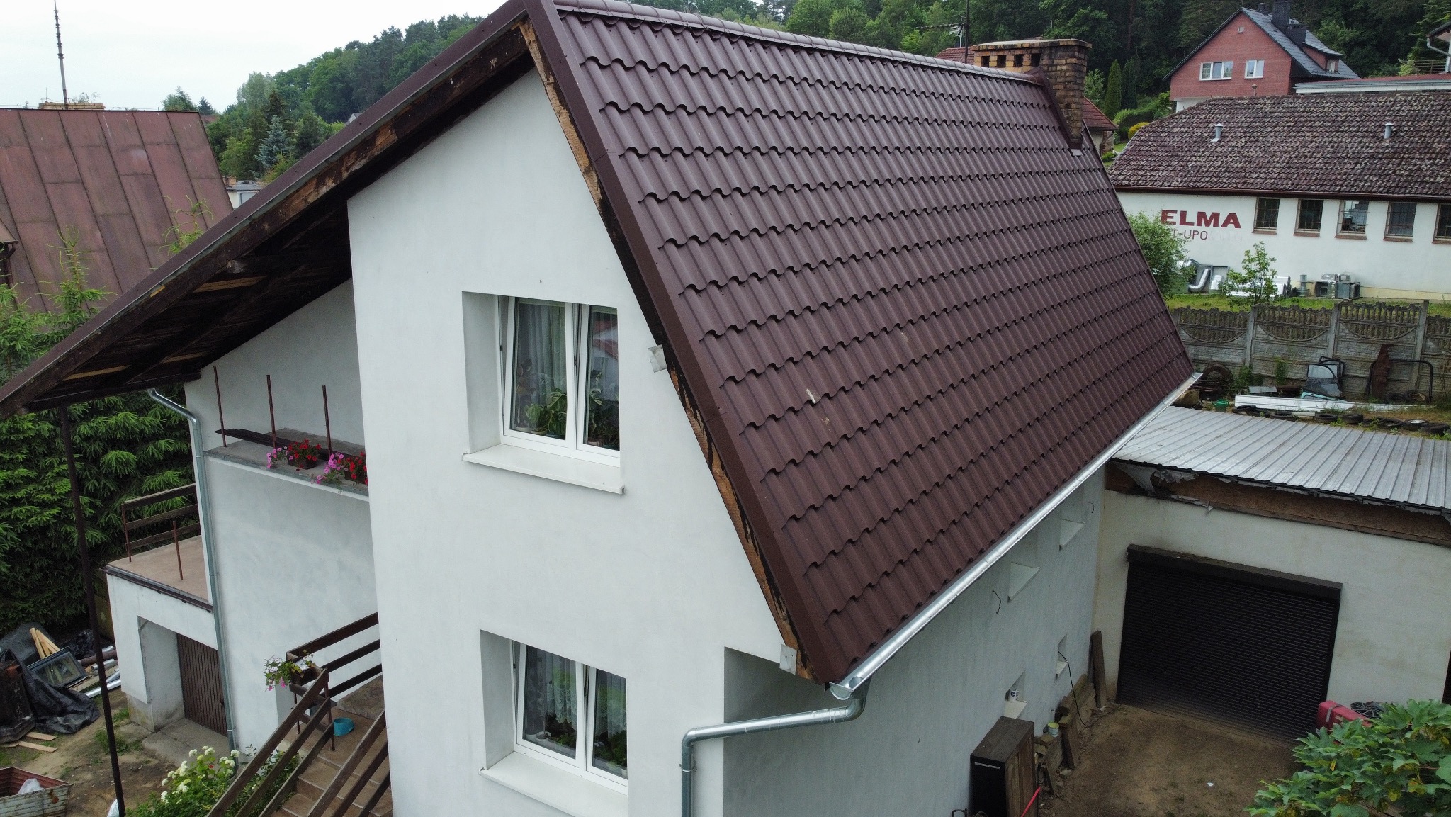 Dom z nowym, brązowym dachem z blachodachówki, widoczny z góry. Rynny zamontowane, okna białe. W tle zieleń i zabudowa jednorodzinna.