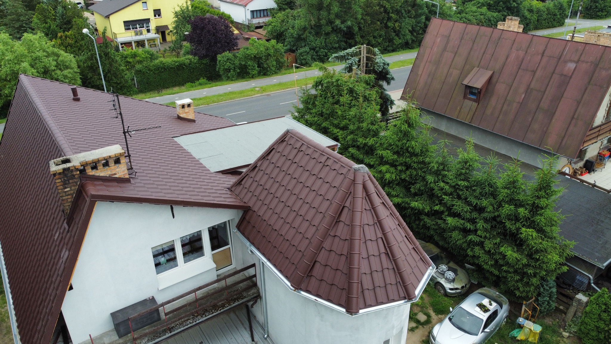 Brązowy dach z dachówki ceramicznej na domu z białymi ścianami, widziany z góry. W tle zieleń drzew i zabudowa mieszkalna. Widoczne rynny i komin.