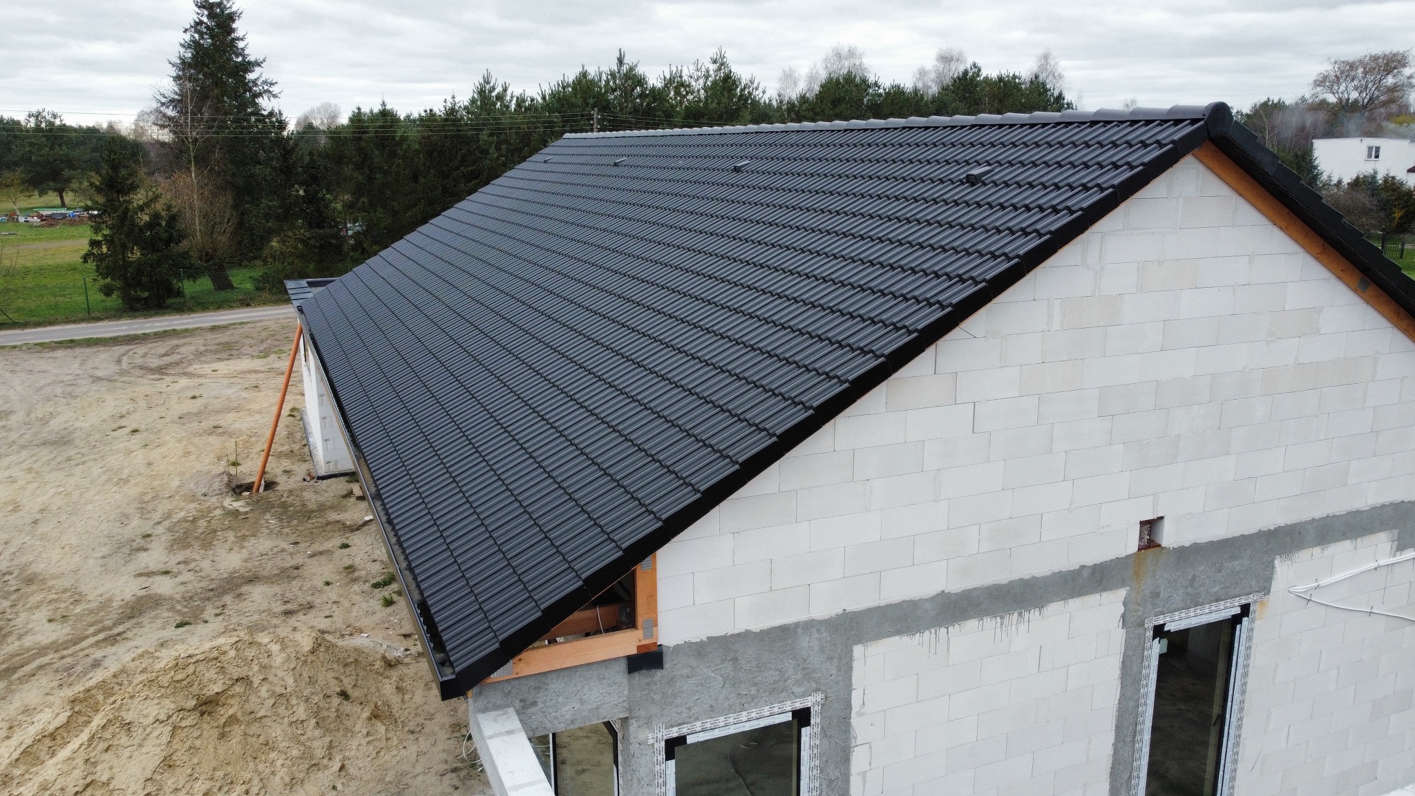Czarny dach z dachówki ceramicznej na surowym budynku z białego bloczka, widok z góry, w tle drzewa. Dachówka ułożona równo, widoczne detale łączeń.