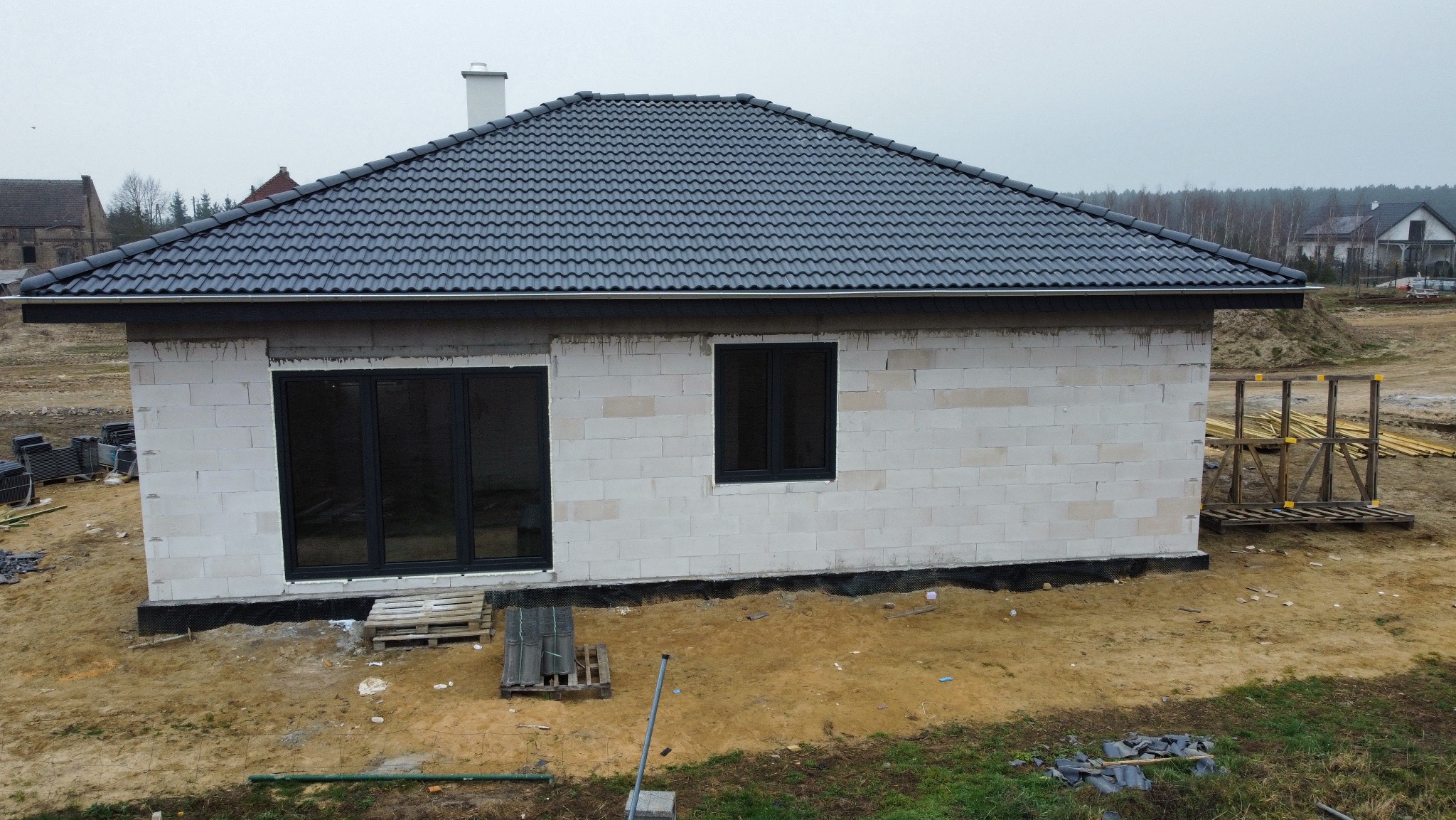 Dom w trakcie budowy z ciemnoszarą dachówką ceramiczną. Widoczne białe ściany z betonu komórkowego, czarne okna i przygotowane drewniane elementy konstrukcyjne.