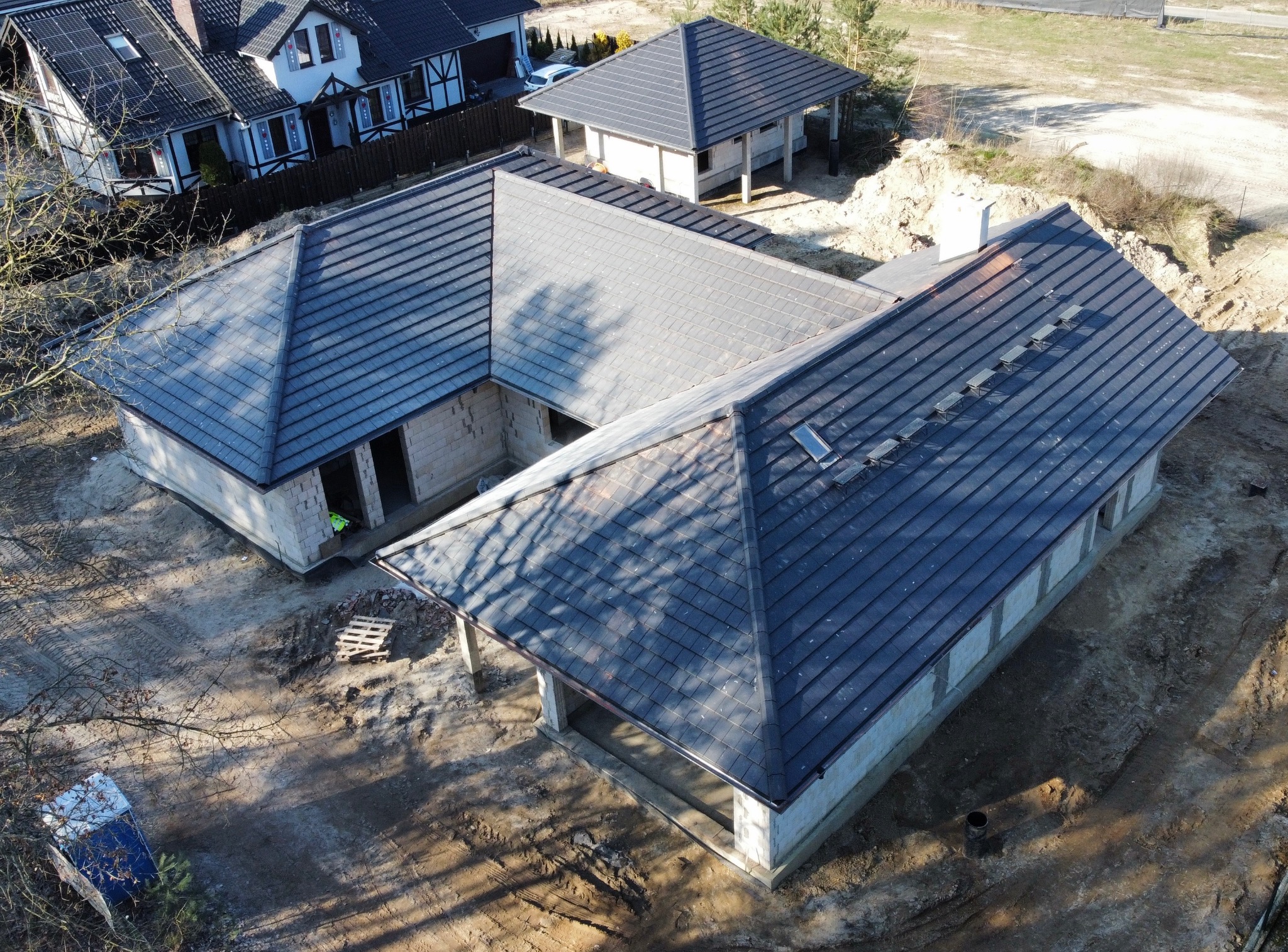 Ukończony dach z ciemnej dachówki ceramicznej na domu w kształcie litery U, widok z lotu ptaka. W tle drugi budynek z identycznym dachem i zabudowa jednorodzinna.