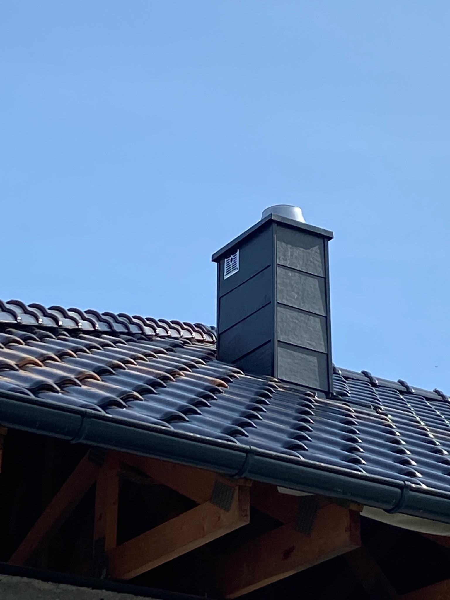 Czarny, kwadratowy komin z metalowym daszkiem wystający z ciemnobrązowego dachu krytego dachówką ceramiczną, na tle błękitnego nieba.
