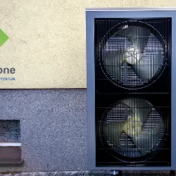 Szara pompa ciepła marki Nesheat z dwoma wentylatorami, umieszczona na zewnątrz budynku, obok okna piwnicznego i logo firmy Green Zone z napisem 'Pompy ciepła Klimatyzacja'.