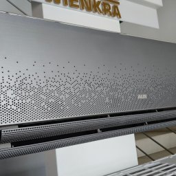 Green Zone - Szary klimatyzator AUX z perforowanym panelem przednim, widoczne żaluzje wylotowe powietrza, zamontowany w pomieszczeniu z dekoracją świąteczną w tle.