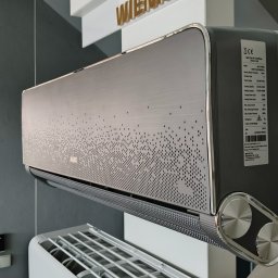Green Zone - Nowoczesny klimatyzator AUX w kolorze srebrno-grafitowym z dekoracyjnym wzorem perforacji na panelu przednim, widoczna tabliczka znamionowa z parametrami technicznymi, tło stanowi szara ściana...