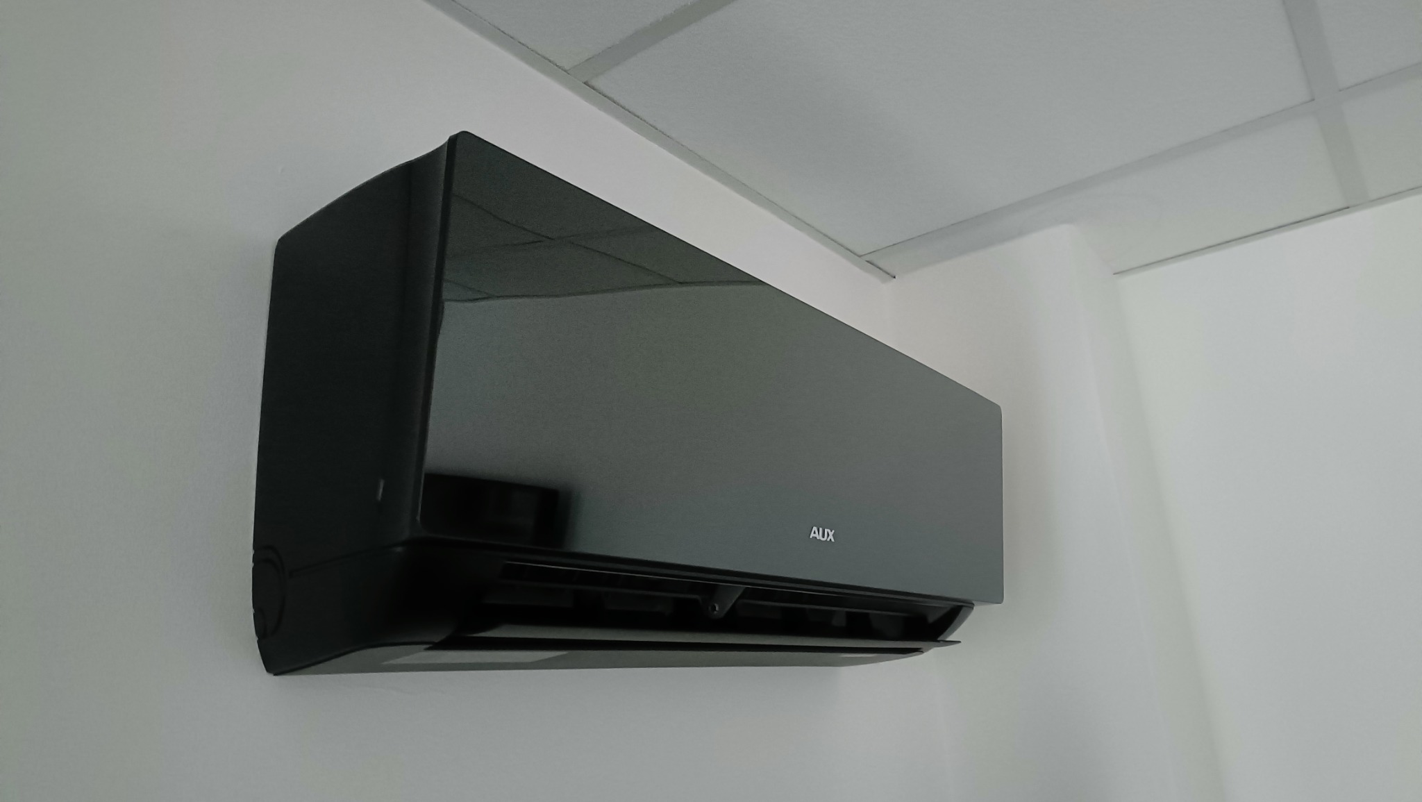 Czarny klimatyzator marki AUX zamontowany na białej ścianie w pomieszczeniu z kasetonowym sufitem.