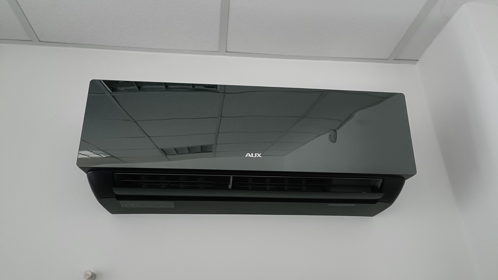 Czarny klimatyzator AUX zamontowany na białej ścianie w pomieszczeniu z kasetonowym sufitem, widoczne odbicie sufitu w obudowie klimatyzatora.