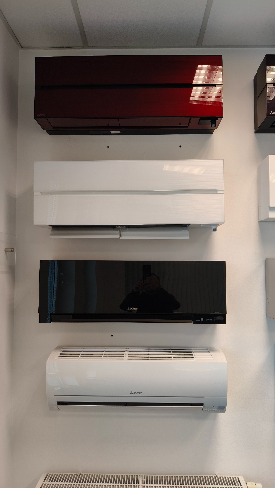 Wystawa czterech klimatyzatorów Mitsubishi Electric w różnych kolorach (czerwony, biały, czarny, biały) zamontowanych na ścianie, od góry do dołu.