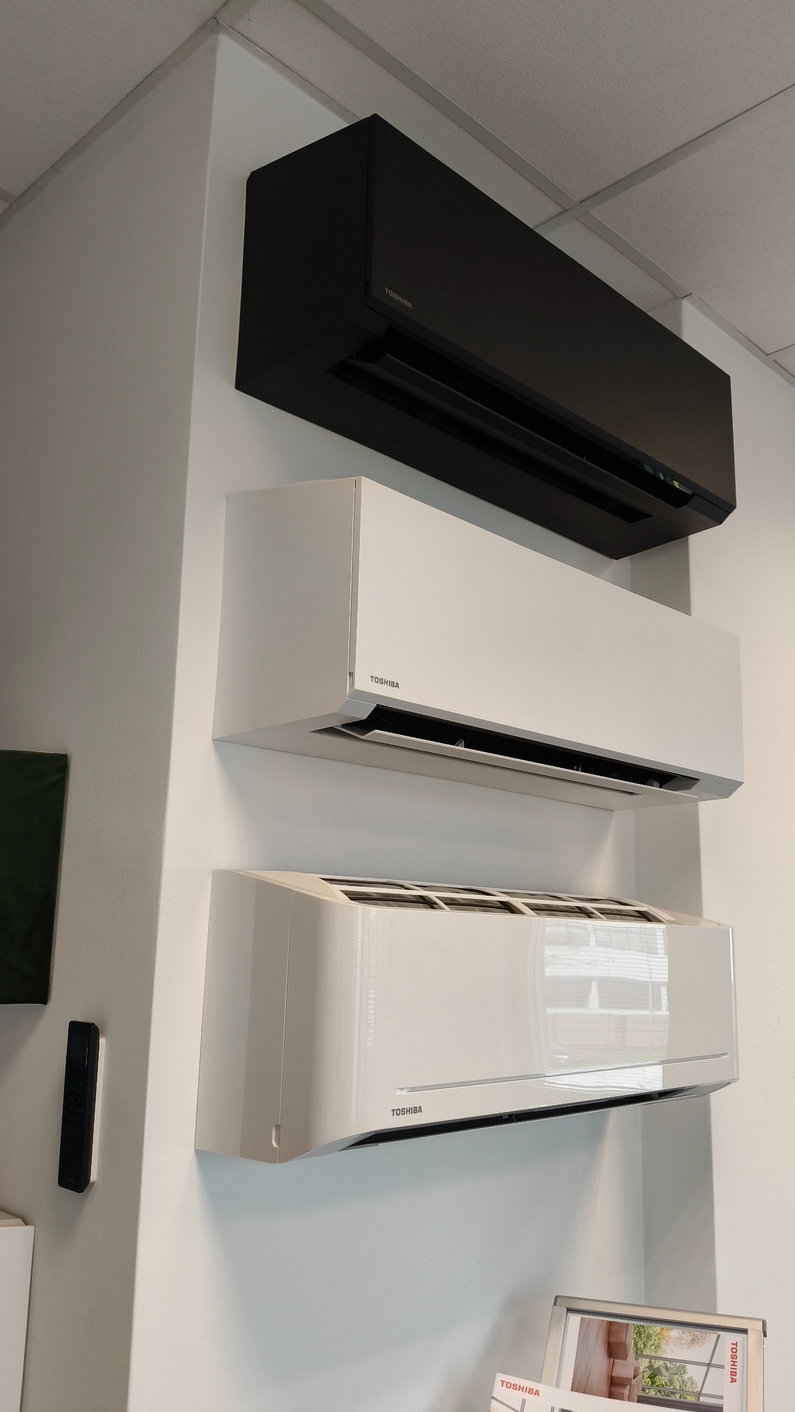 Ekspozycja trzech klimatyzatorów Toshiba w kolorach czarnym i białym, zamontowanych na białej ścianie w salonie sprzedaży.