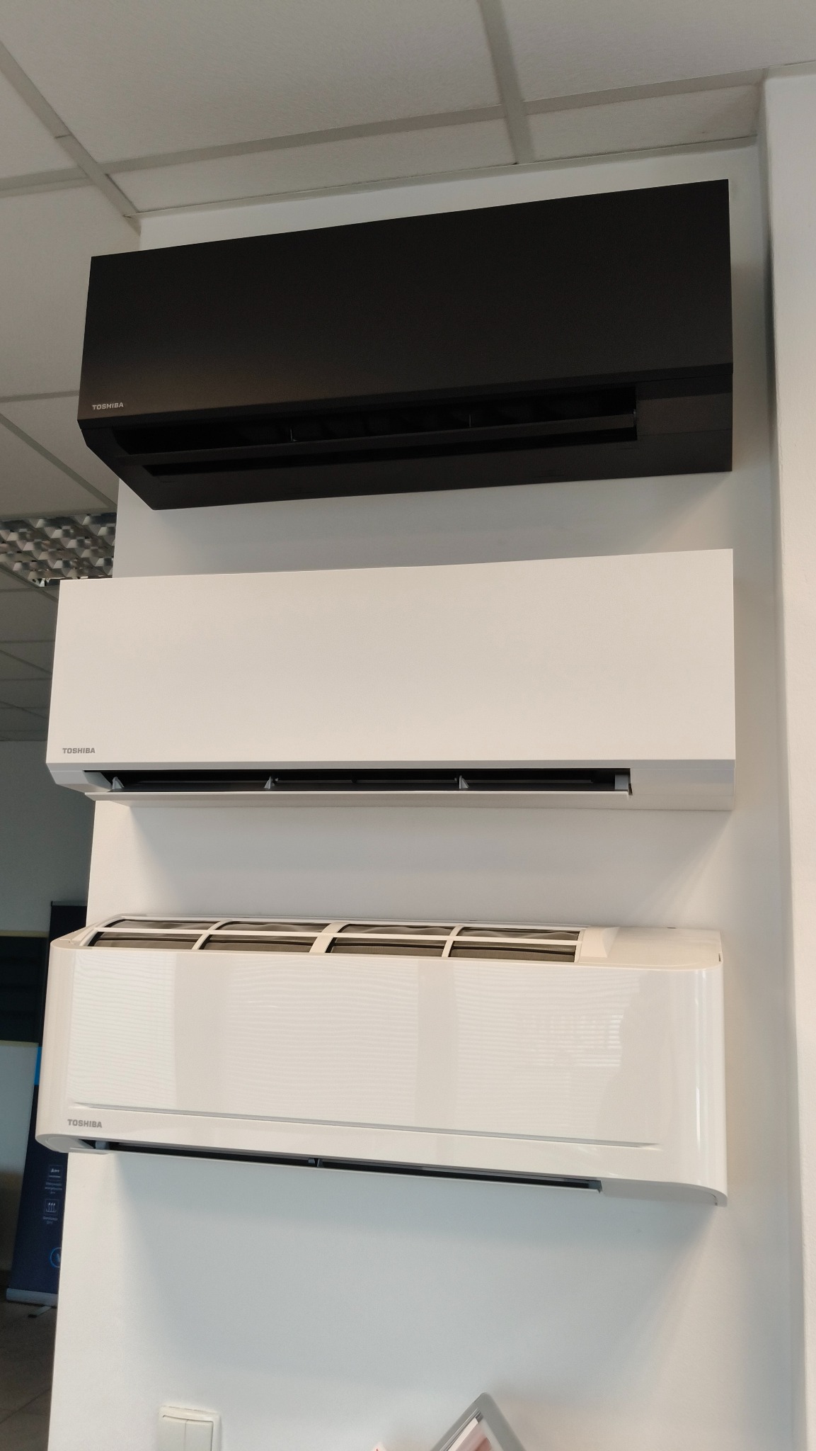 Trzy jednostki klimatyzacyjne Toshiba zamontowane na białej ścianie, od góry: czarna, biała i biała z widocznymi filtrami.