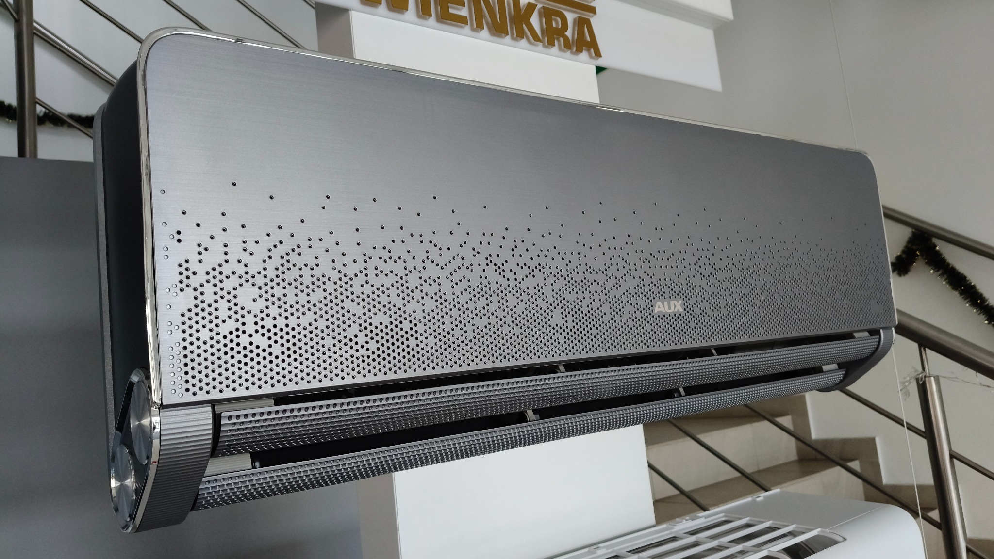 Szary klimatyzator AUX z perforowanym panelem przednim, widoczne żaluzje wylotowe powietrza, zamontowany w pomieszczeniu z dekoracją świąteczną w tle.