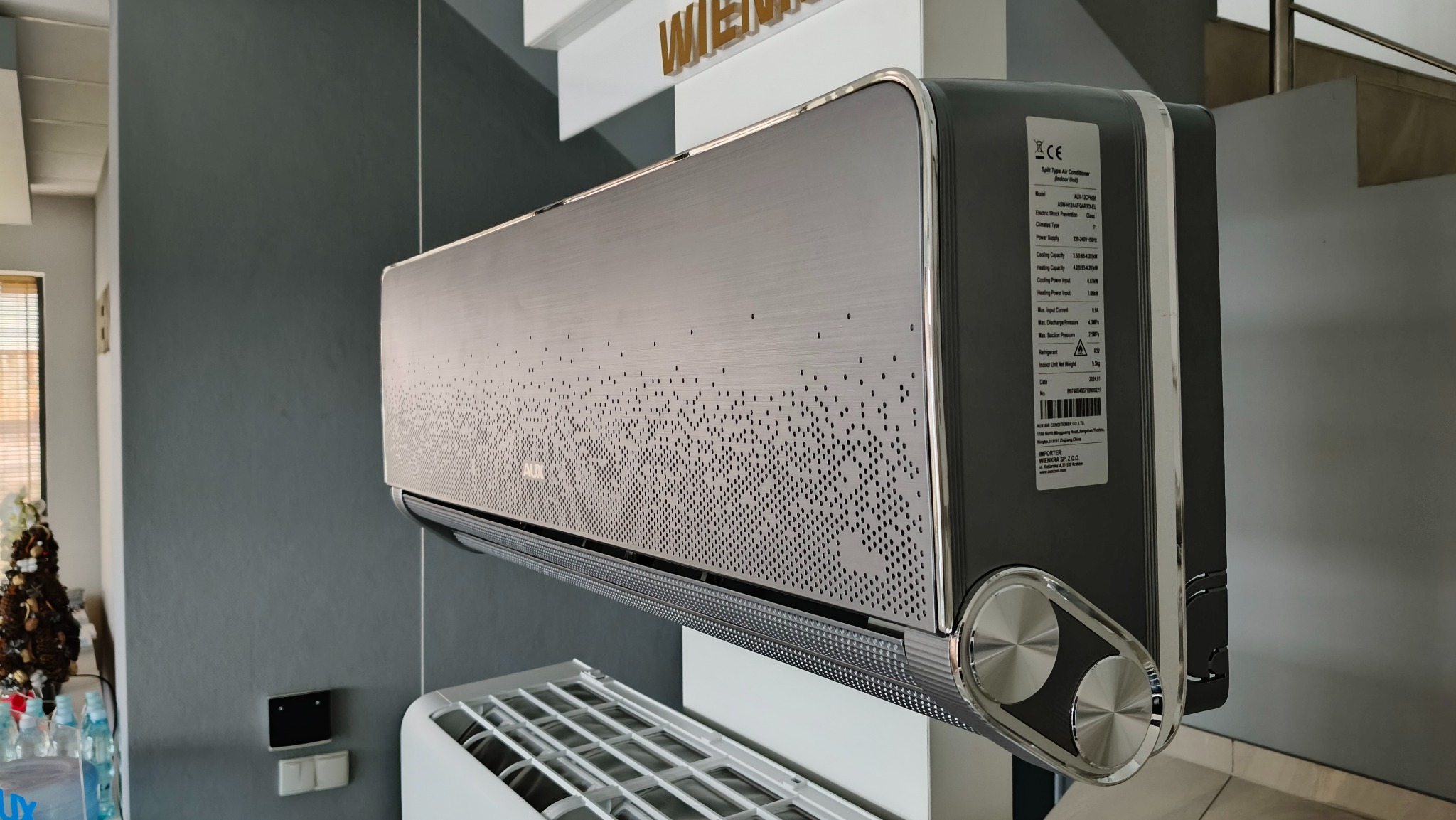 Nowoczesny klimatyzator AUX w kolorze srebrno-grafitowym z dekoracyjnym wzorem perforacji na panelu przednim, widoczna tabliczka znamionowa z parametrami technicznymi, tło stanowi szara ściana...
