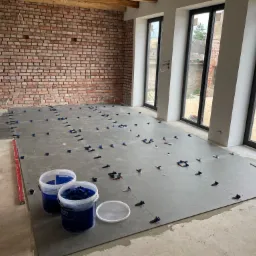 Układanie szarych płytek podłogowych z systemem poziomowania, widoczne dwa wiadra z elementami systemu, ceglana ściana i duże okna w tle.