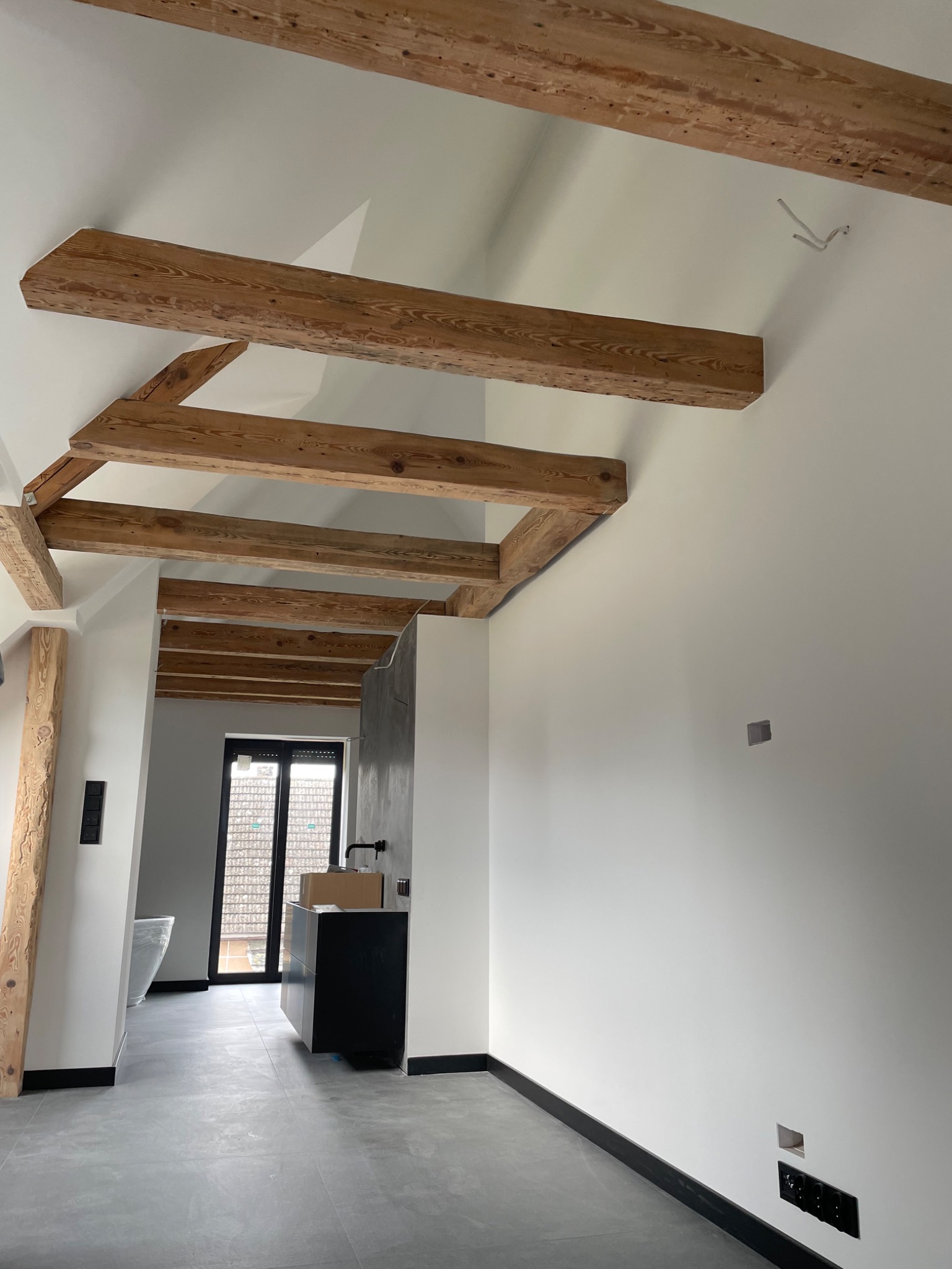 Wnętrze w trakcie prac wykończeniowych: surowe drewniane belki stropowe, świeżo pomalowane białe ściany, szara podłoga z czarnymi listwami przypodłogowymi, widoczna łazienka w głębi i czarna szafka...