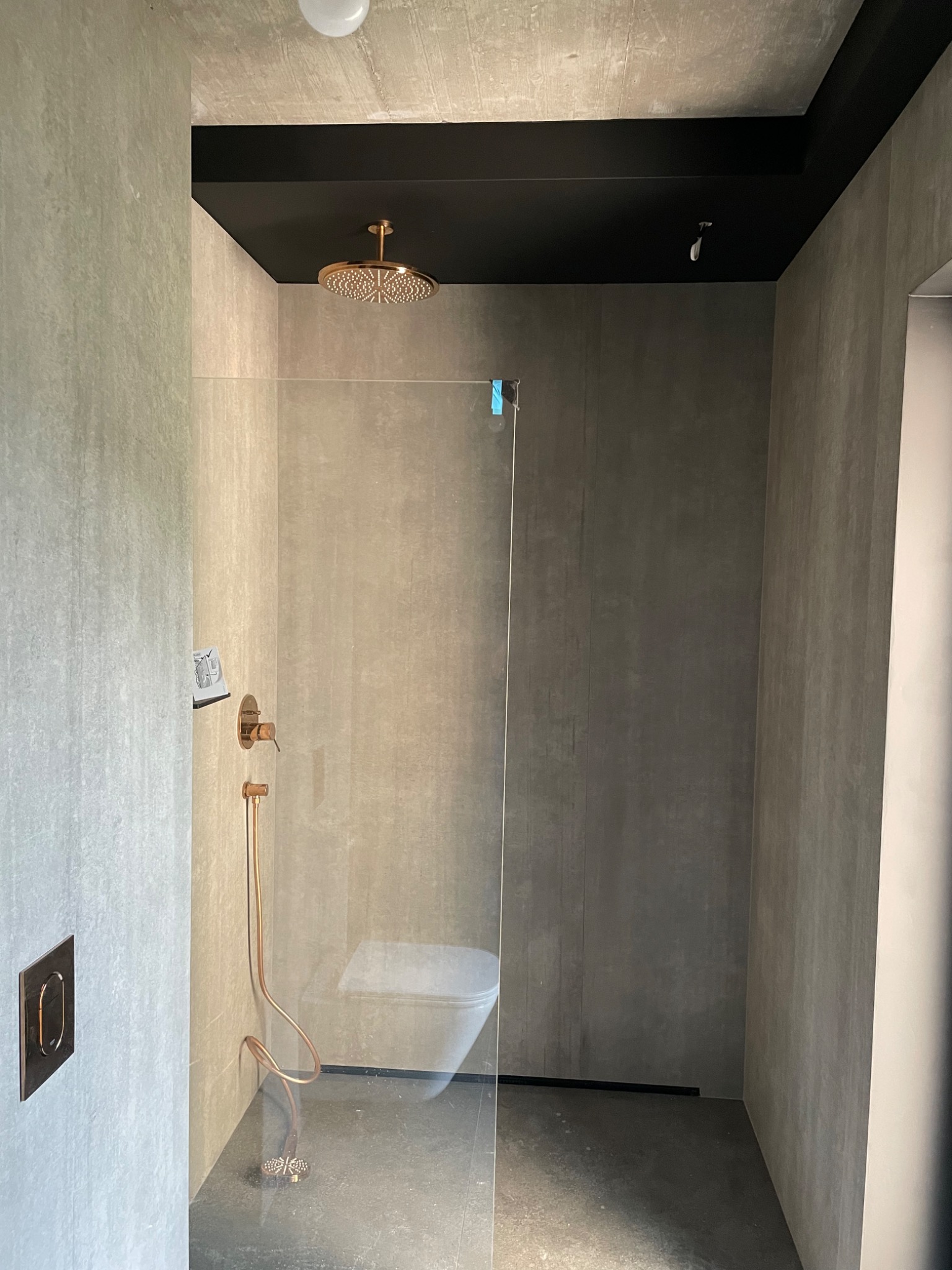 Nowoczesna łazienka z kabiną prysznicową typu walk-in, wykończona płytkami imitującymi beton, z miedzianą armaturą i czarnym sufitem podwieszanym.